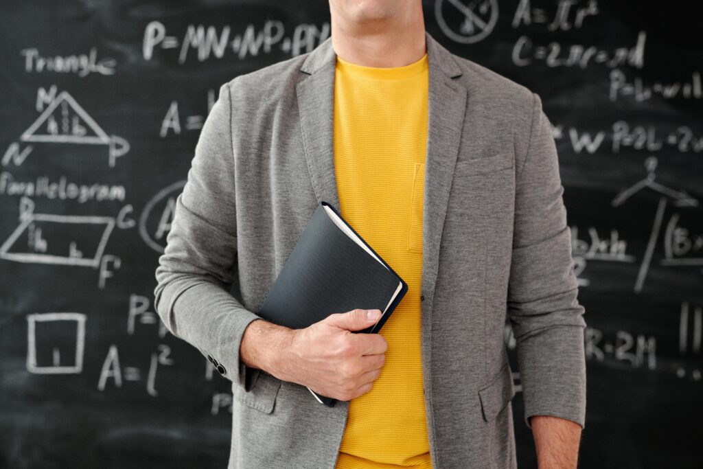 Teacher holding a book in front of a chalkboard with writing on it.